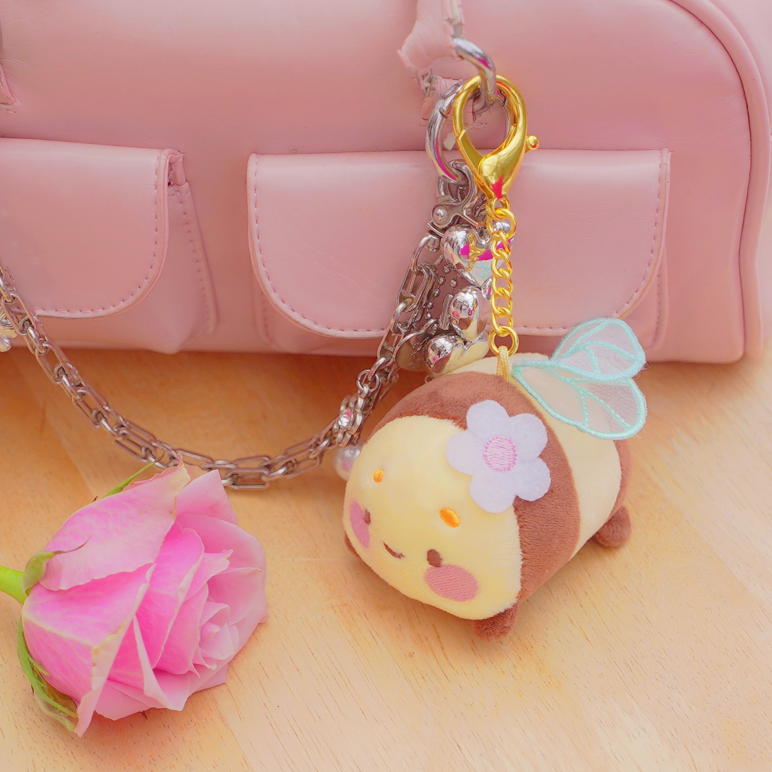 A cute plush bee keychain charm with a white flower on its head, light blue wings, and a gold clasp, shown hanging from a silver chain next to a pink rose and a pink handbag on a wooden surface.
