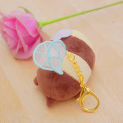 A small plush keychain shaped like a brown and cream striped character with light blue wings and a gold clasp, photographed on a light wood surface next to a pink rose.