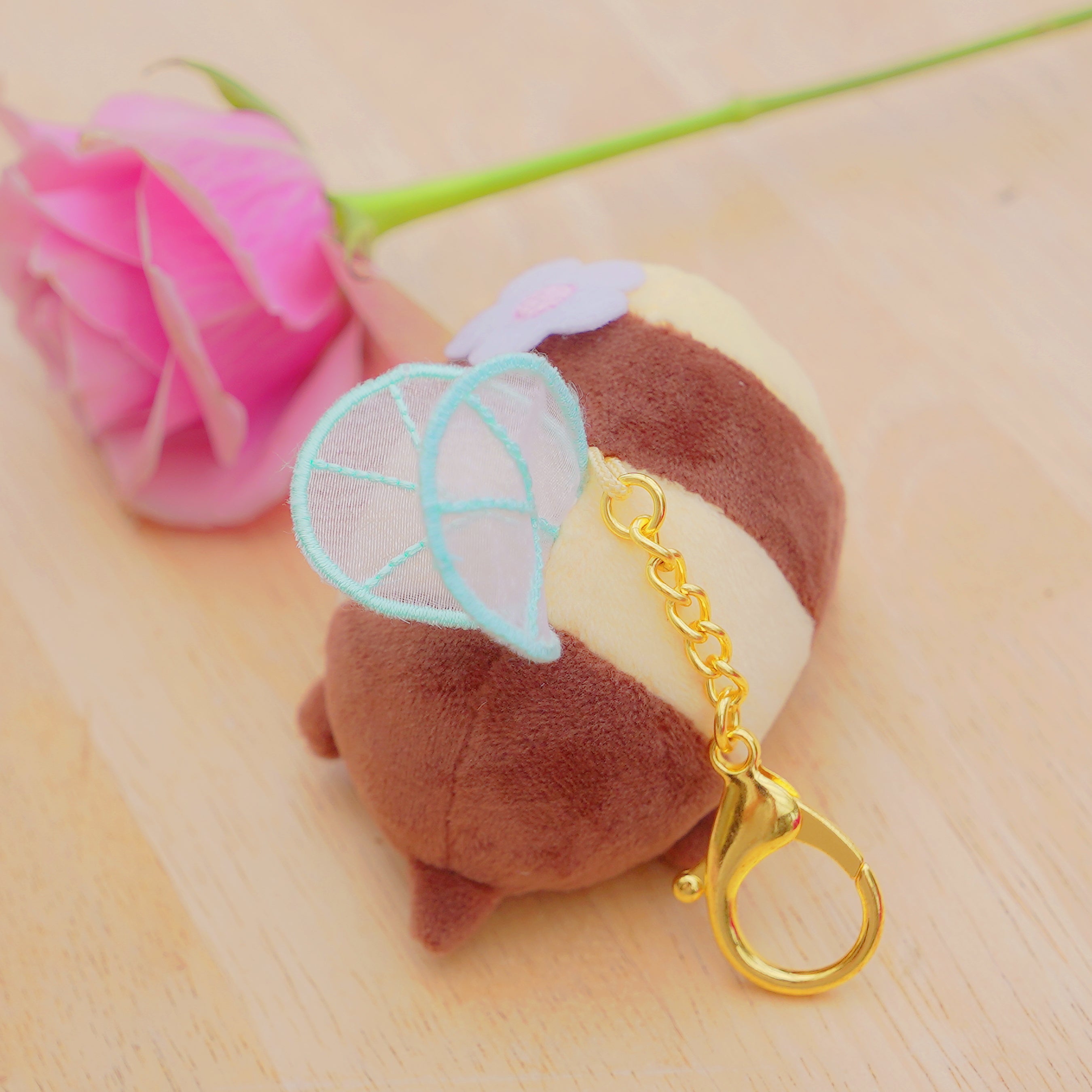 A small plush keychain shaped like a brown and cream striped character with light blue wings and a gold clasp, photographed on a light wood surface next to a pink rose.