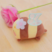 A small plush bee toy with brown and cream stripes, a light blue translucent wing, and a lavender flower applique, placed on a light wood surface next to a pink rose stem.