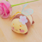 A cute plush bee keychain with embroidered wings, a flower on its head, and a gold key ring, resting on a light wooden surface next to a pink rose.
