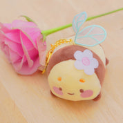 A cute plush keychain shaped like a small character with a yellow face, brown hair, a white flower, and light blue wings, resting on a light wood surface next to a pink rose stem.