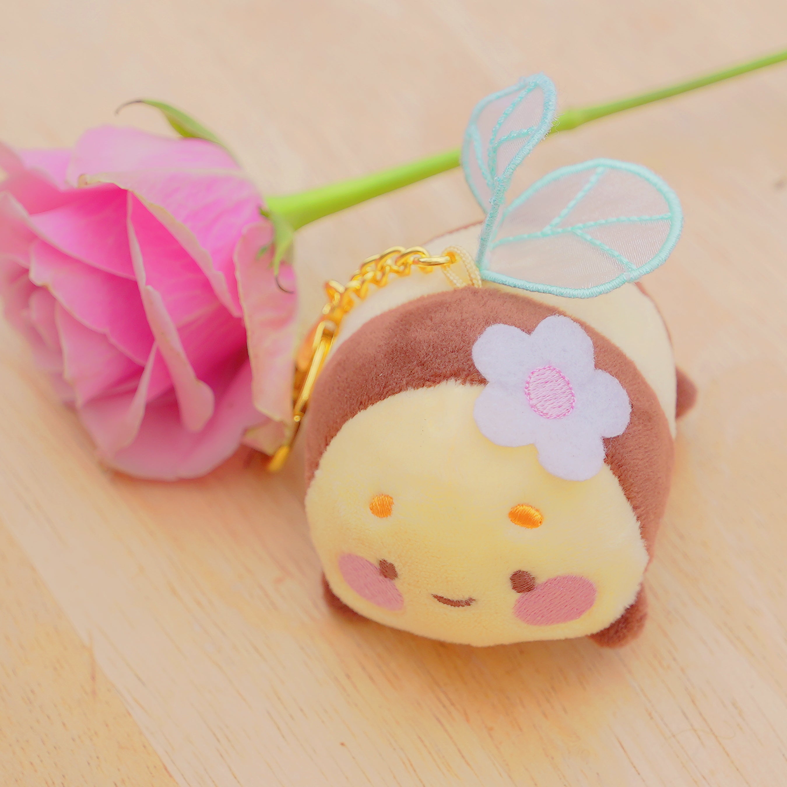 A cute plush keychain shaped like a small character with a yellow face, brown hair, a white flower, and light blue wings, resting on a light wood surface next to a pink rose stem.