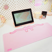 A large, pastel pink coquette bow gaming desk pad displayed on a desk with a laptop and smartphone.