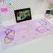 A large, rectangular gaming desk pad with colorful, cartoon-style space-themed characters, displayed on a desk with a tablet and a phone.